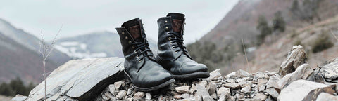 A pair of black leather lace-up Floris van Bommel hiking boots resting on a rocky mountain path with a blurred mountain landscape in the background.