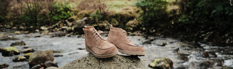 A pair of light brown suede Floris van Bommel ankle boots resting on a rock in a shallow outdoor stream, with a blurred natural forest background.