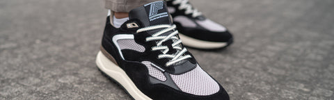 A close-up of a Floris van Bommel black and white sneaker being worn, with a second matching shoe blurred in the background on a gray pavement surface.