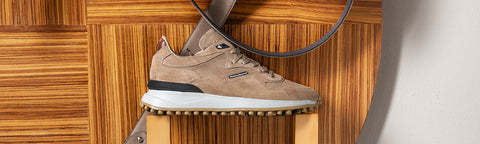 A single low-top Floris van Bommel sneaker in taupe suede and mesh with a white midsole and black heel accent, displayed on a wooden riser against a wood-panelled background.