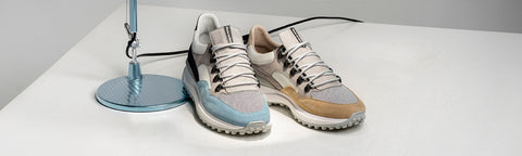 A pair of low-top athletic Floris van Bommel sneakers with a light grey and beige upper, white laces, and a light blue heel accent, displayed on a white surface next to a blue lamp.