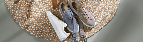 Three casual low-top Floris van Bommel sneakers arranged on a woven round mat: a white sneaker, a light blue sneaker, and a taupe-beige sneaker.