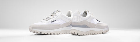 A pair of low-top Floris van Bommel sneakers in white and light grey, featuring textured soles and side stripes, displayed against a clean light grey studio background.