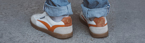 A close-up of a person's feet wearing white Floris van Bommel sneakers with orange side stripes and gum soles, paired with cuffed blue jeans, standing on a grey concrete surface.