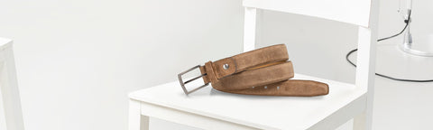 A brown leather belt brand van bommel with a silver metal buckle, resting on a white wooden chair in a minimalist room setting.