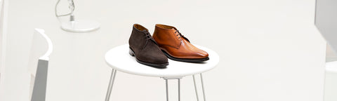 A pair of men's van Bommel lace-up dress shoes, one dark brown and one tan/cognac, displayed side-by-side on a small white round table in a bright, minimalist interior.
