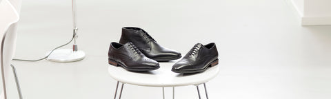 Three black leather Floris van Bommel shoes arranged on a small white round table in a bright, minimalist room with a white floor and wall.