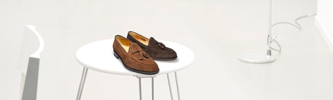 A pair of brown suede tassel van Bommel loafers displayed on a small white round table in a minimalist white room setting.