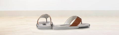A pair of minimalist thong sandals brand floris van bommel with light grey soles and straps, featuring a brown leather strap accent, displayed on a light wooden surface against a white background.
