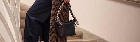 A person wearing dark blue wide-leg pants and a brown plaid skirt carries a black leather Floris van Bommel shoulder bag with a patterned scarf strap while walking up an indoor staircase.