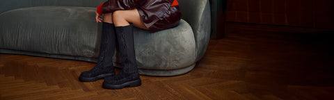 A person seated on a grey upholstered bench in a room with wood flooring, wearing black knee-high Floris van Bommel boots.