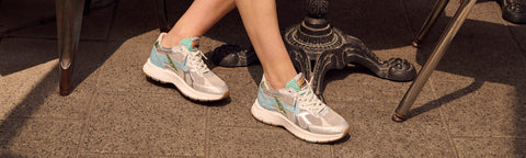 A person wearing light grey and mint green Floris van Bommel sneakers, seated outdoors on a stone floor near a metal table base.
