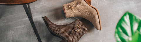 Two Floris van Bommel ankle boots with a snakeskin pattern, one in brown and one in beige, are displayed on a gray concrete floor next to a green leaf.