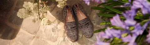 A pair of women's Floris van Bommel slip-on loafers with a brown animal print pattern, styled on a stone surface with white flowers and purple flowers in the foreground.