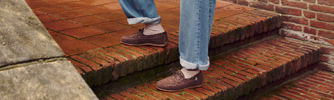 A person wearing light blue jeans and brown Floris van Bommel lace-up loafers walks up red brick stairs outdoors.