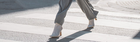A person walking across a crosswalk outdoors, shown from the knees down, wearing light grey wide-leg trousers and Floris van Bommel ankle boots with heels.