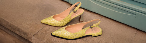 A pair of Floris van Bommel slingback heels in a textured yellow material, resting on a stone step next to a light blue door.