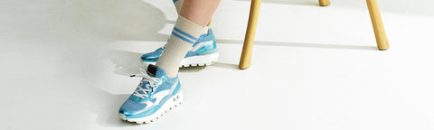 A close-up of a person's feet wearing light blue and white Floris van Bommel sneakers with white socks, standing on a white floor next to the legs of a wooden stool.