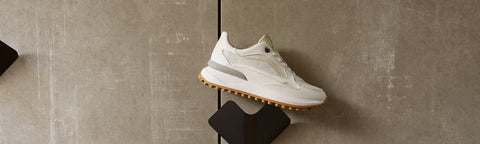 A white Floris van Bommel sneaker with light grey overlays and a gum sole, displayed on a black stand against a textured grey concrete wall.