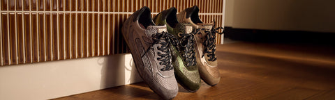 Three Floris van Bommel sneakers in grey, olive green, and beige tones leaning against a slatted wall on a wooden floor.