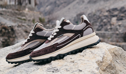 A pair of Floris van Bommel sneakers in brown, beige, and grey suede and mesh, resting on a large rock with a blurred rocky mountain background.