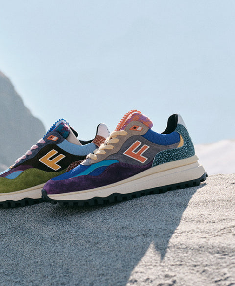 A pair of colorful Floris van Bommel sneakers with a multicolour suede and mesh upper, featuring a chunky white midsole and black outsole, set against a blurred mountain background.