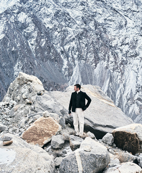 Floris van Bommel wearing a black jacket and off-white trousers, stands on a rocky mountain slope with a large snow-dusted mountain face in the background.