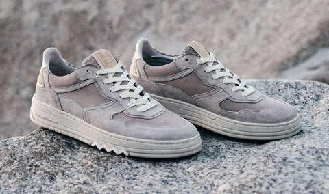 A pair of Floris van Bommel sneakers in a muted taupe-grey suede and leather mix, resting on a textured grey stone surface with a blurred natural rock background.