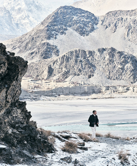 Floris van Bommel in a dark blazer and white trousers, stands on a snowy mountain path with rugged rocky peaks and a frozen lake in the background.