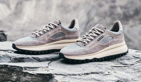 A pair of Floris van Bommel sneakers in a muted taupe-grey suede and mesh material, resting on a grey stone surface with a blurred natural rocky background.