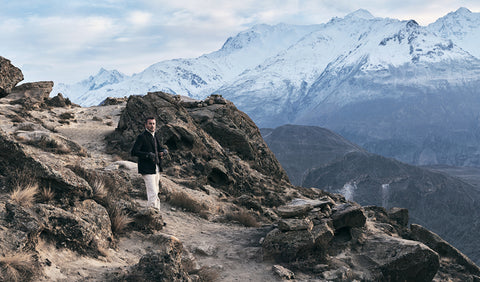 Floris van Bommel stands on a rocky mountain path with snow-capped peaks in the background, wearing a black jacket and light-colored pants.