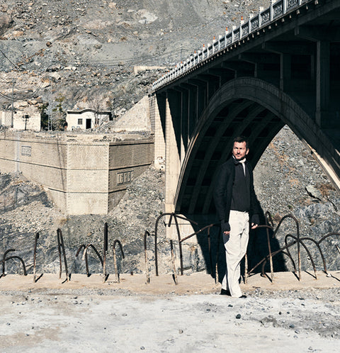 Floris van Bommel in a black jacket, black shirt, and light beige trousers, stands under a large concrete arch bridge in a rocky, arid outdoor setting.