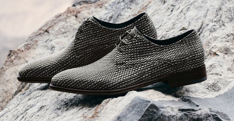 A pair of men's Floris van Bommel lace-up dress shoes with a textured, woven snakeskin-like pattern in black and brown, resting on a natural grey rock surface.