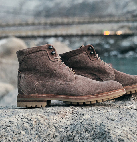 A pair of Floris van Bommel brown suede ankle boots with rugged lug soles, resting on a rocky surface with a blurred outdoor background.