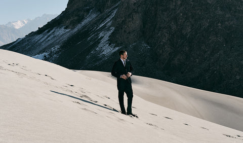 Floris van Bommel in a dark suit stands on a vast sand dune with a dark rocky mountain slope behind him and distant snow-capped mountains in the background.