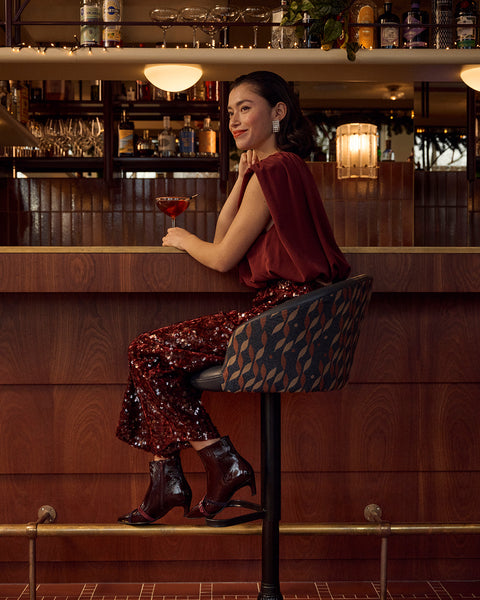A woman sits on a patterned bar stool at a wooden bar, wearing a burgundy velvet top and matching sequined burgundy pants with dark Floris van Bommel heeled boots, holding a glass of red drink.