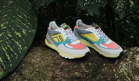 A pair of Floris van Bommel sneakers resting on a tree trunk outdoors, surrounded by large green leaves. The shoes feature a mix of light grey, yellow, pink, teal, and white panels.