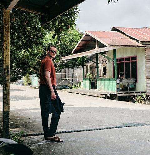 Floris van Bommel wearing a rust-orange short-sleeve polo shirt, dark trousers, and brown sandals stands on a concrete path in front of a rustic house with a corrugated metal roof and green trim. He is holding a dark jacket under one arm.