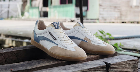 A pair of Floris van Bommel Classic Running sneakers in beige, white, and blue, resting on a rustic wooden surface with an outdoor background.
