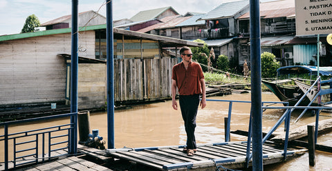 Floris van Bommel wearing a rust-orange short-sleeve shirt and dark trousers, walks across a wooden dock over murky water, with rustic houses and boats in the background.