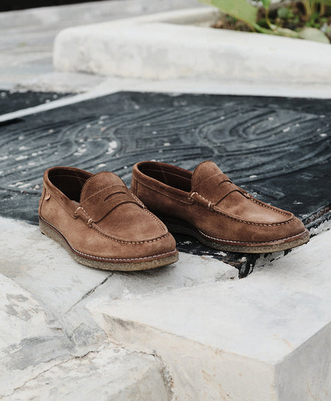 A pair of brown suede Floris van Bommel loafers resting on a black textured mat set against a light stone background.