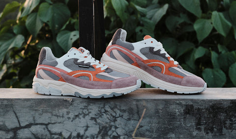 A pair of Floris van Bommel sneakers in dusty mauve, grey, white, and orange, resting on a weathered stone ledge with green foliage in the background.