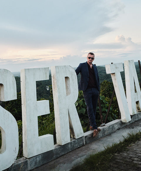 Floris van Bommel, wearing a dark navy blazer, black shirt, and sunglasses, stands in front of large white letters.