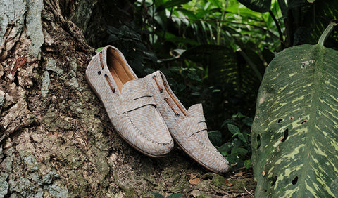 A pair of light beige Floris van Bommel leather loafers with a subtle snakeskin texture, resting on a tree trunk in a lush green outdoor setting.