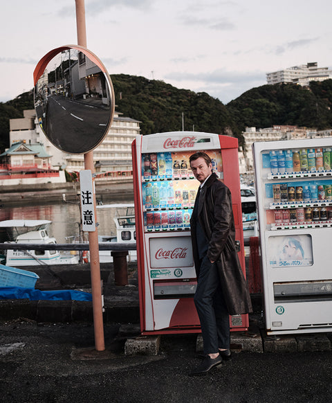 Floris van Bommel in a dark coat and trousers stands beside a red Coca-Cola vending machine in an outdoor harbor setting, with another vending machine, a round traffic mirror, boats, and hills in the background.