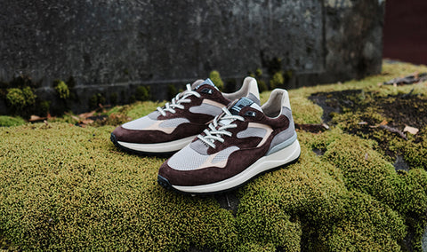 A pair of Floris van Bommel sneakers in brown, grey, and off-white, styled on a bed of green moss outdoors against a dark stone wall.