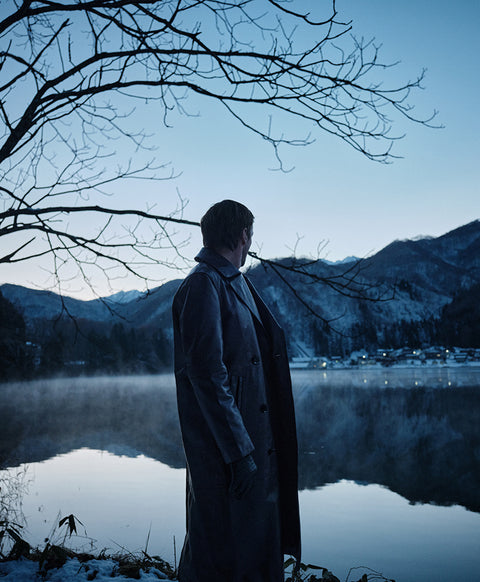 Floris van Bommel in a long dark coat stands by a calm lake, with bare tree branches overhead and mountains in the background.