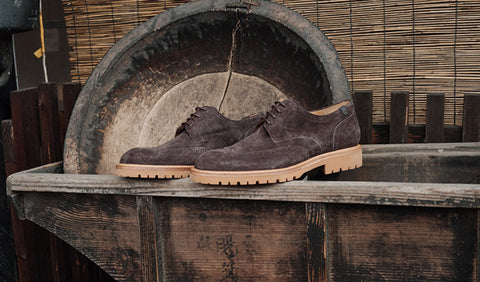 A pair of dark brown suede Floris van Bommel shoes with tan soles, displayed on a rustic wooden crate in front of a large wooden bowl.