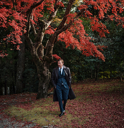 Floris van Bommel in a dark navy blue three-piece suit with a brown leather coat walks under a large tree with vibrant red autumn leaves in a forest.