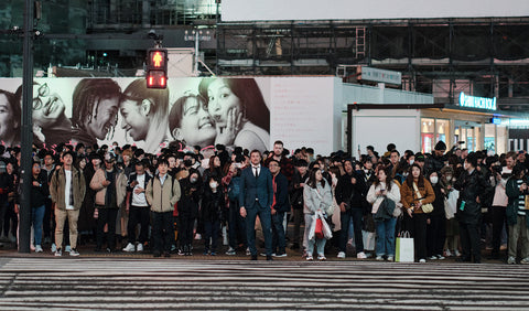 A large crowd of people standing on a city street crosswalk at dusk with in the middle Floris van Bommel, with a giant black-and-white billboard in the background and urban buildings and signage around them.